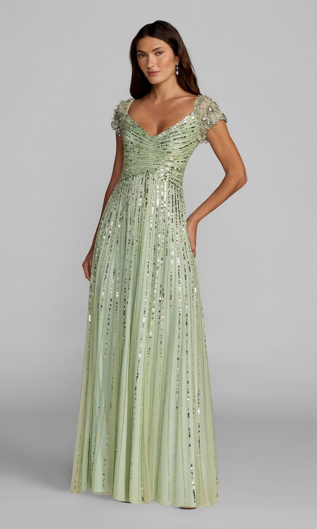 A woman in the Mac Duggal 23042 Short Sleeve Sequin Formal Dress—floor-length, light green, with embellished sleeves and vertical sequins—stands against a gray background, perfect for mothers of the bride or formal sequin events.